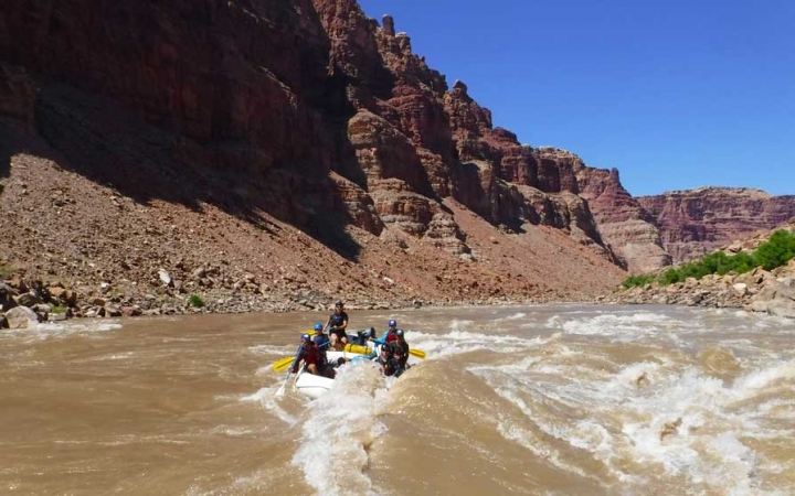 A group of people paddle a raft on whitewater. The river is framed by tall canyon walls.